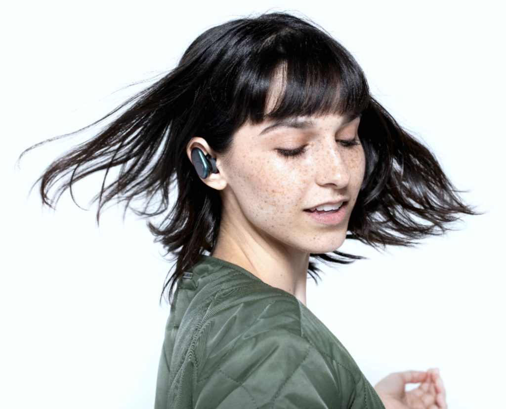 A woman twirls and her hair is in motion, she is highly freckled and wearing a green coat. You can see the SkullCandy Push Ear Buds in her ear. 