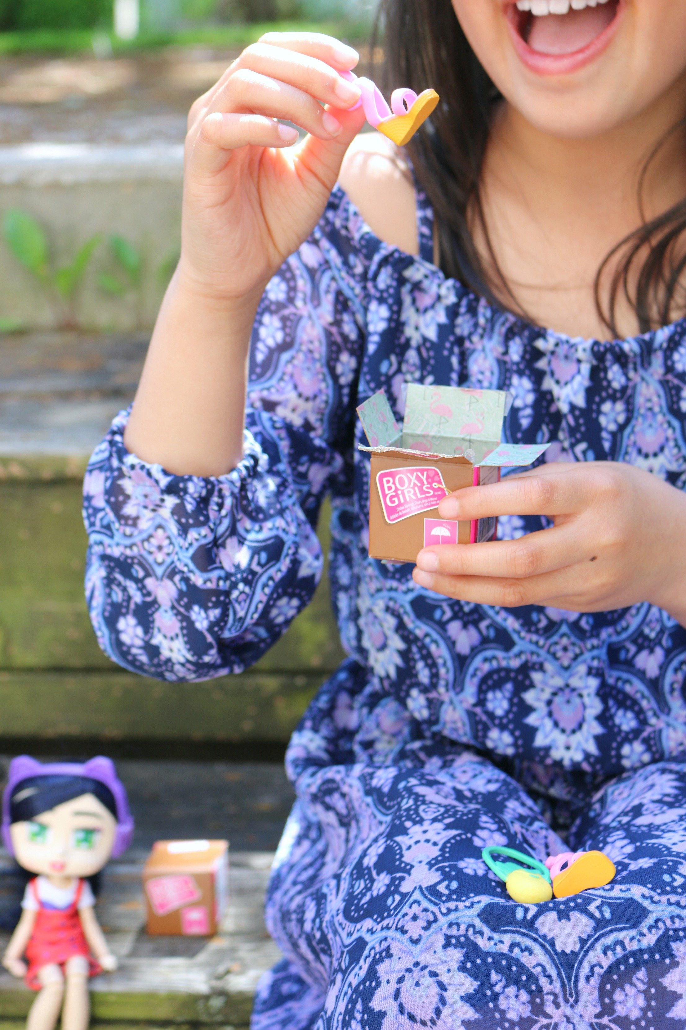 A close up of a little girl unboxing a mini Boxy Girl box, she pulls out a shoe. In the background is a Boxy Girl with another surprise box.