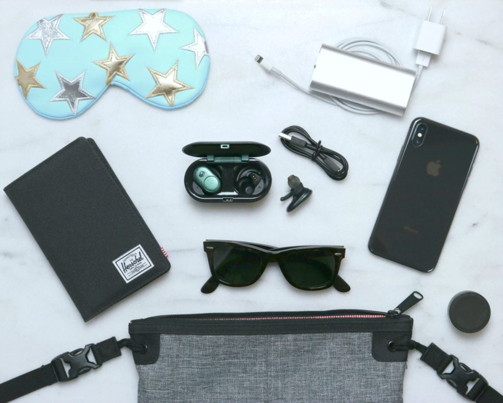 A flat lay picture of a personal messenger bag with the inside components spread out: a passport holder/wallet, sunglasses, Skullcandy Ear Buds, phone, chargers, lip balm, and a sleeping mask. 