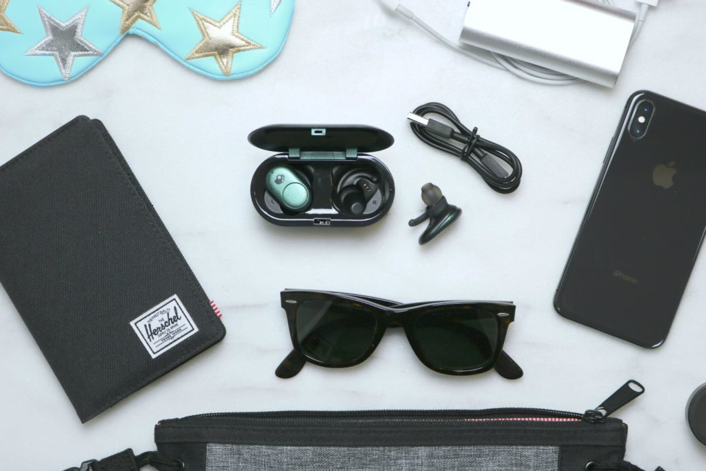 A close up of a flat lay picture of a personal messenger bag with the inside components spread out: a passport holder/wallet, sunglasses, SkullCandy Ear Buds, phone, chargers, lip balm, and a sleeping mask. 