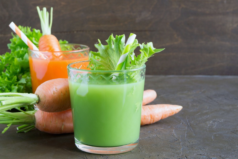 A cup of green juice, carrot juice, surrounded my fresh veggies.