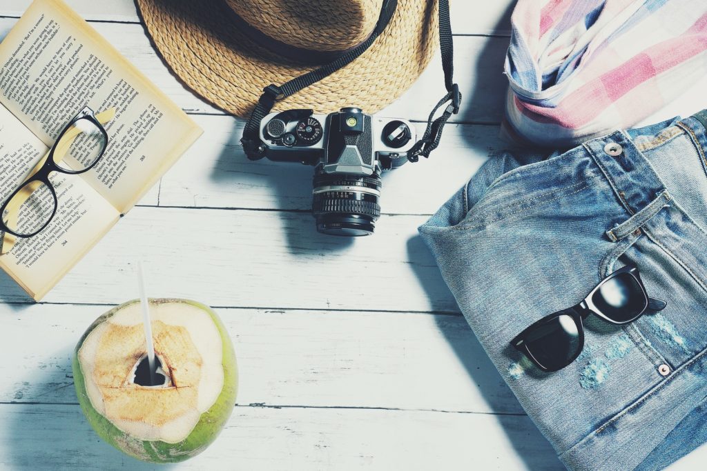 A hat, camera, book with eyeglasses, jeans, a shirt and a coconut drink are shown in flat lay.