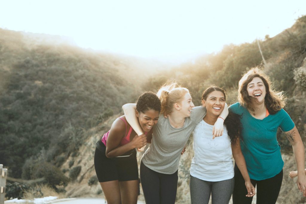 4 mulitcultural friends link arms around necks and laugh on top off a hillside.