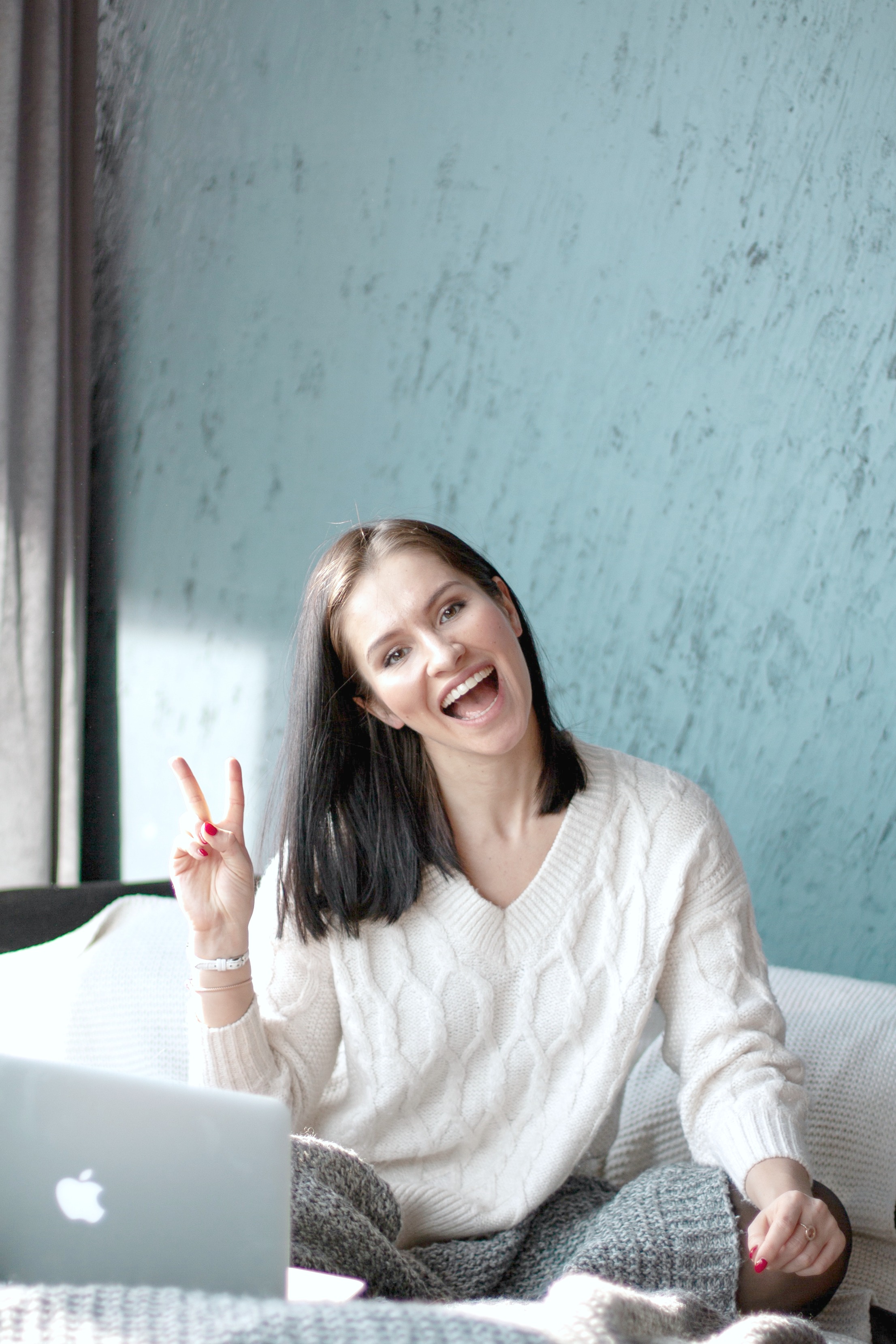 A woman gives a peace sign as she sits happily in bed on her laptop. 
