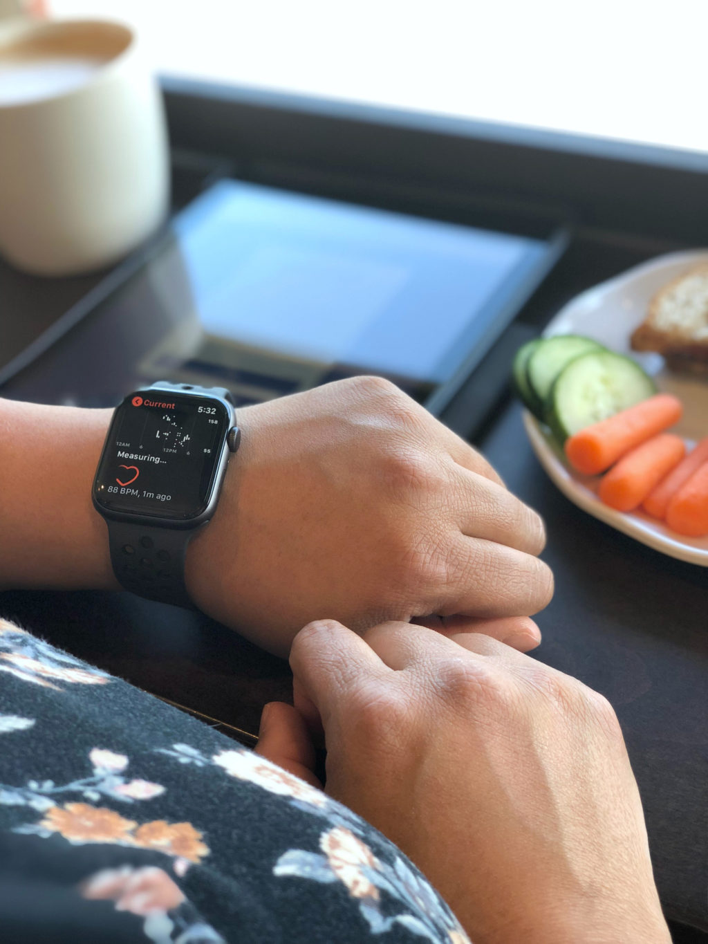 A woman checks her heart rate at a cafe while eating a veggie plate, sandwich, and a latte.