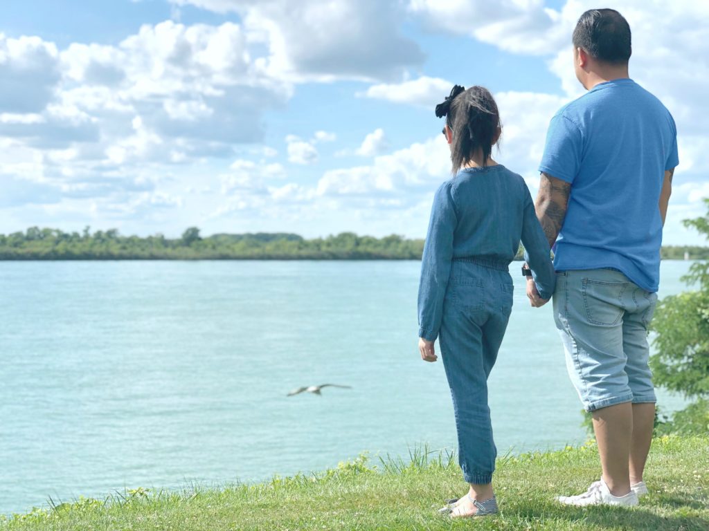 Mimi and Darasak watch a bird fly over the Detroit river while standing on a hillside.