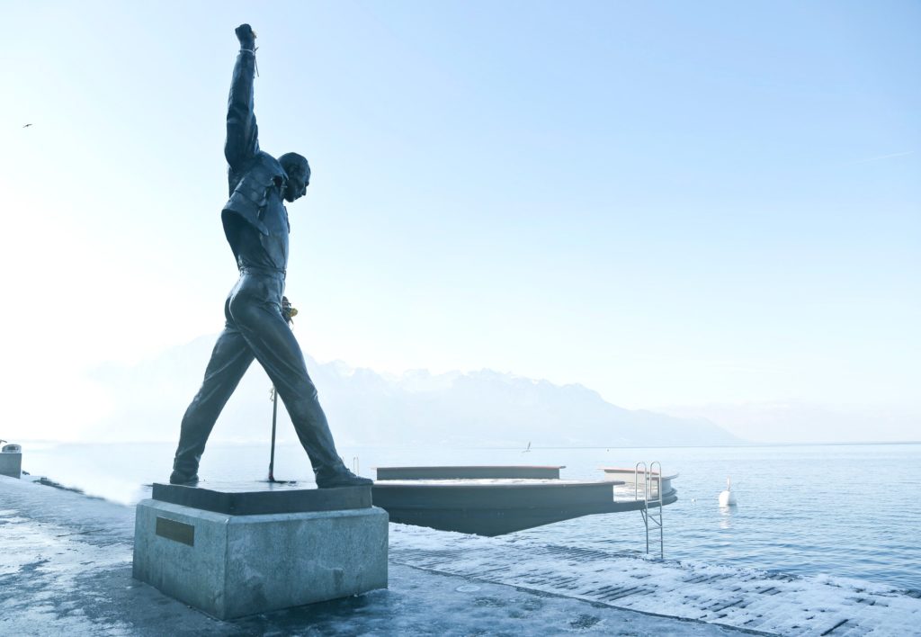 A statue of Freddie Mercury stands in his signature pose alongside a body of water, the sky is behind him.