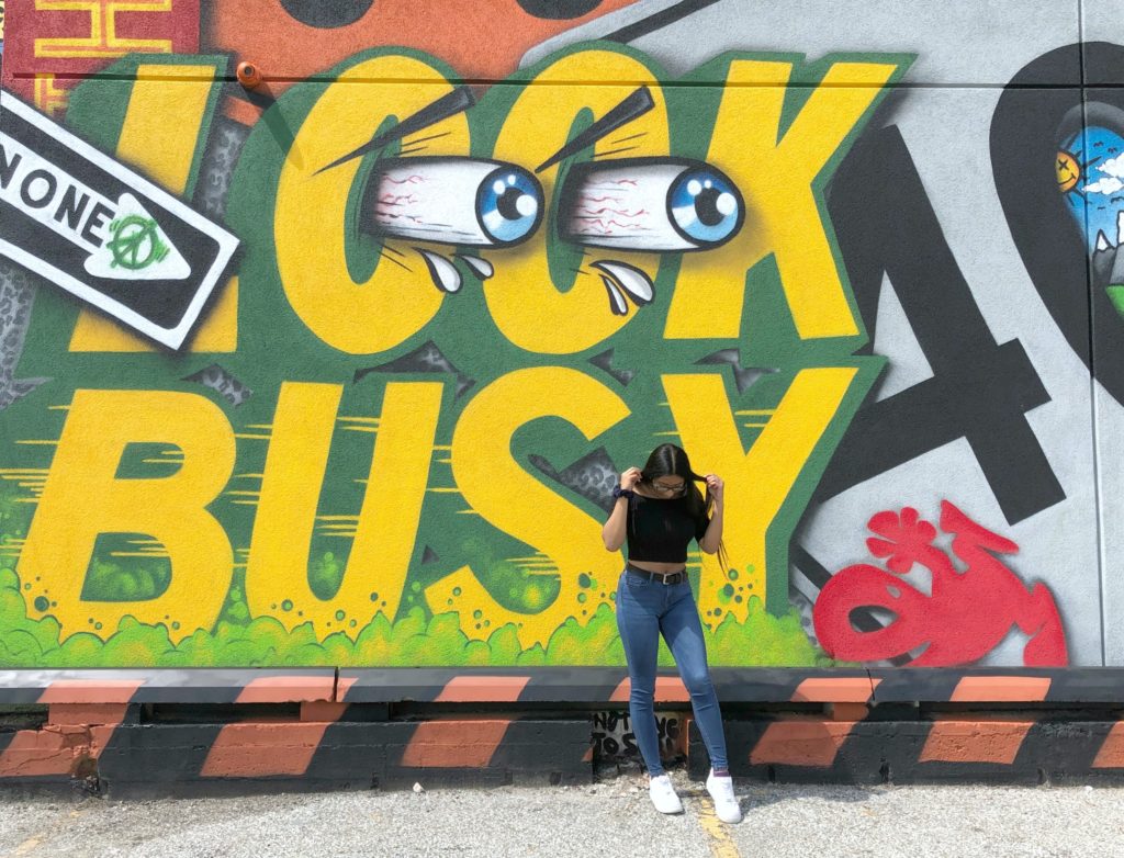 Gabby poses in front of a mural that says 'Look Busy' and looks IG or Snapchat ready.