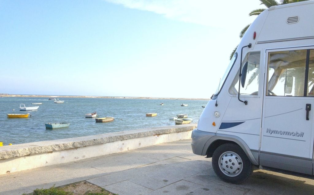 The tip of an RV is shown parked next to a waterfront that is lined with many boats. 