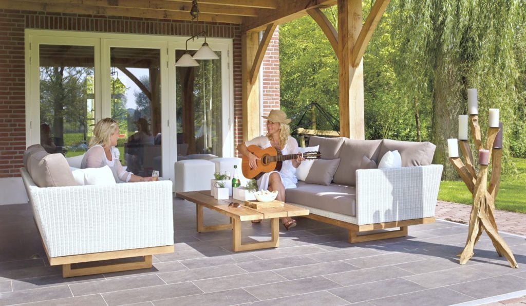 Two women sitting on a patio, the patio has lovely rattan sofas and two women sit on each sofa. One is playing guitar.