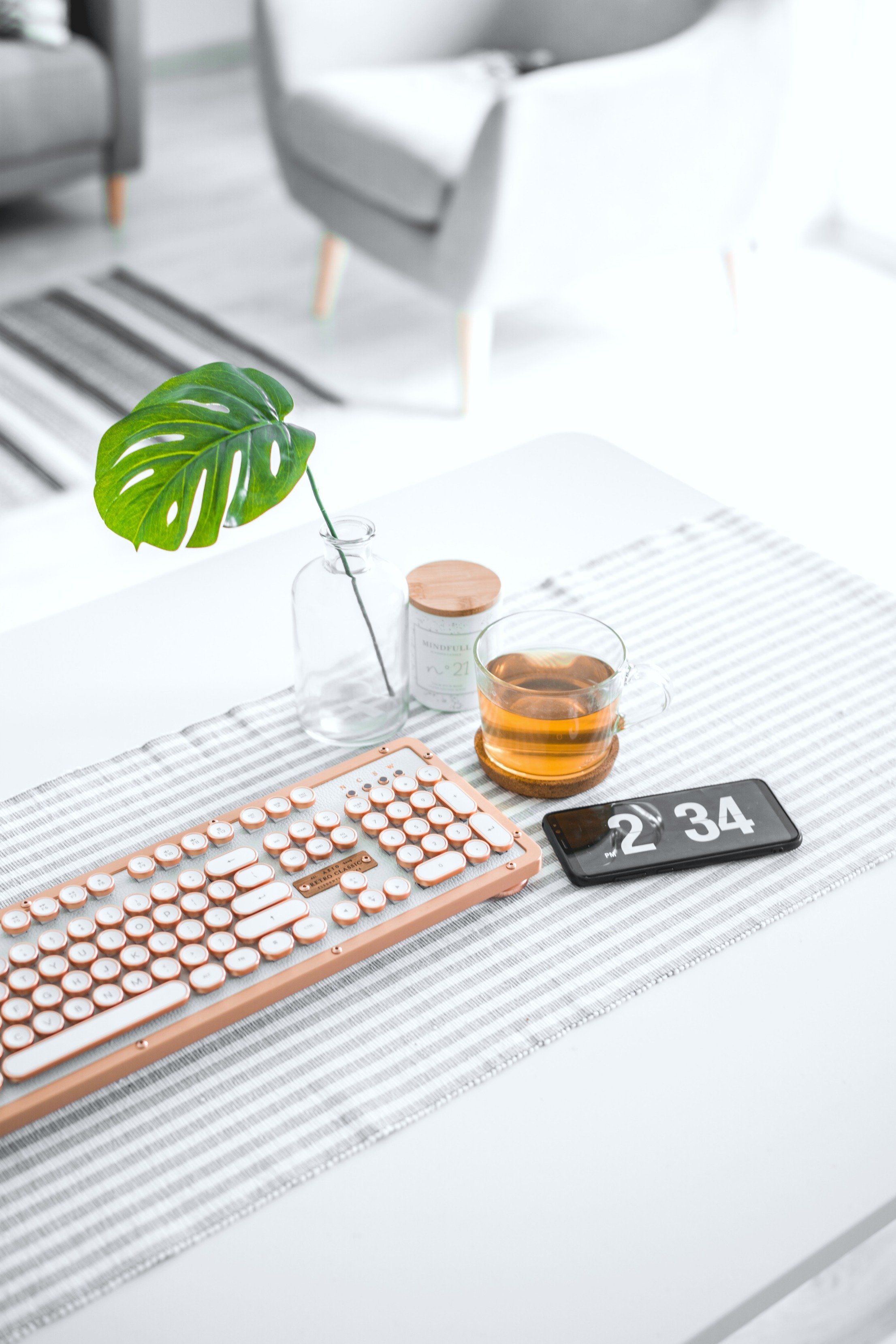 A desk with a plant, a cup of tea, keyboard, and phone are seen. A living room is in the background. 