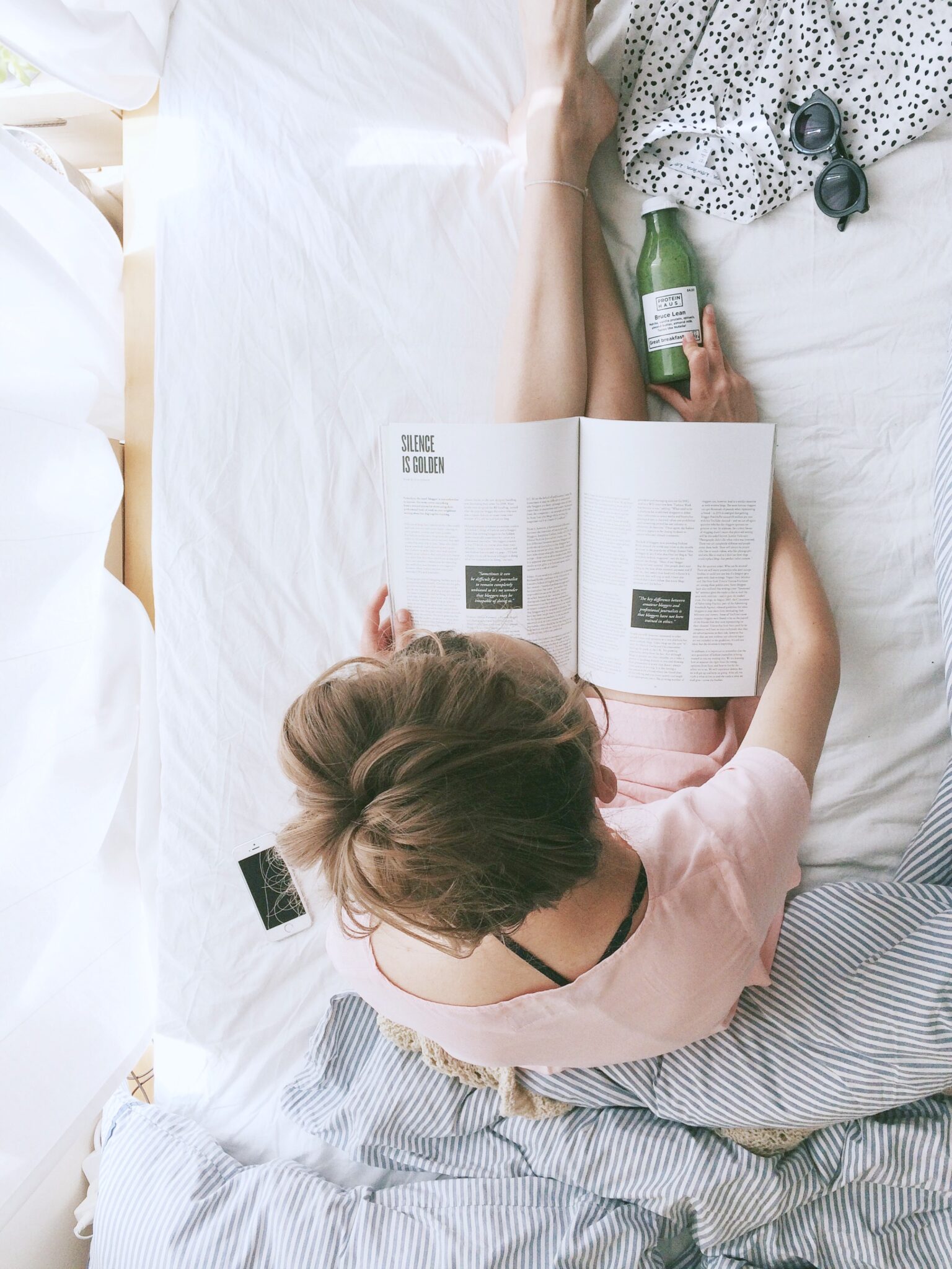 A woman reads a book on her bed, the view is from above and she is holding a green drink in her other hand. 