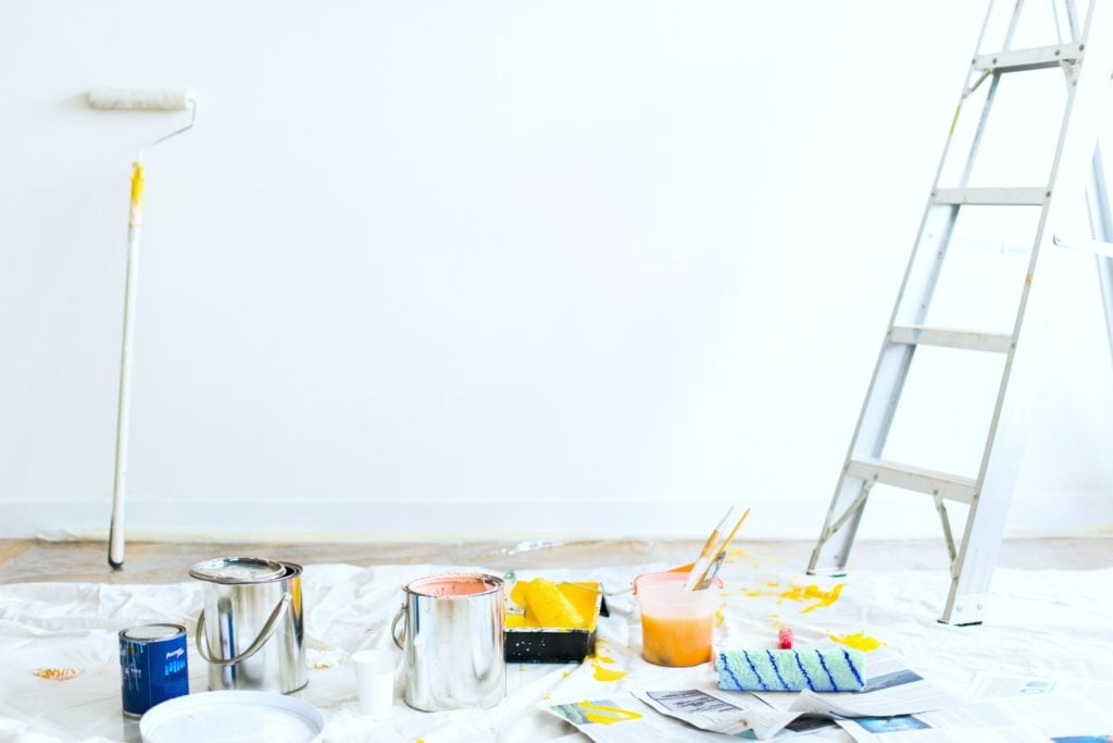 A blank white wall has a paint brush on a pole leaning against it. There is paint materials on the ground on top of a tarp and a ladder is near by. 