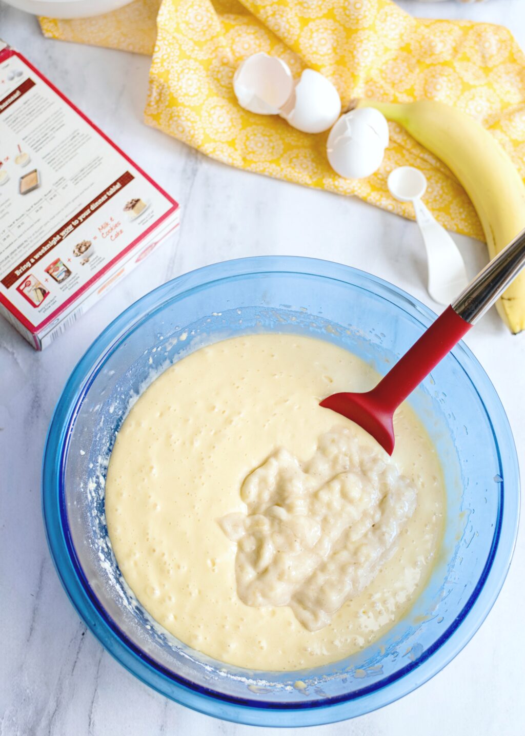 Banana being mixed into cake batter with red spoon.