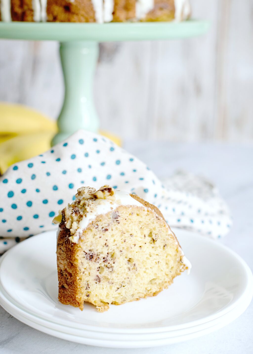 A piece of banana bundt cake cut on a white plate. A cake pedestal is view behind it, along with a polka dot napkin.