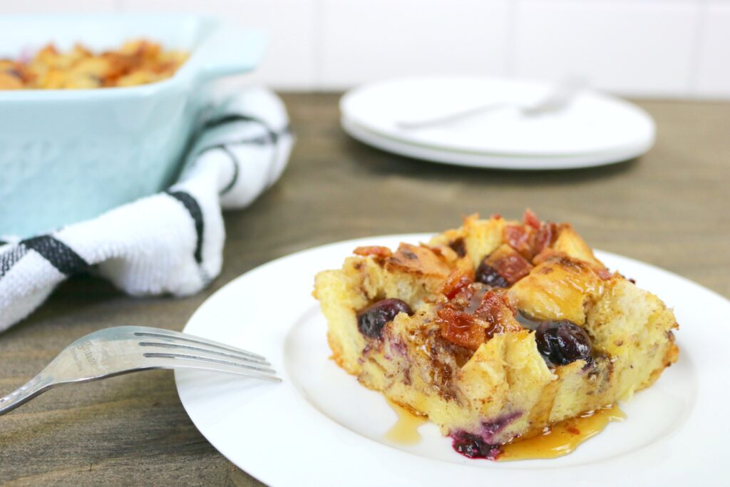 A pan of Blueberry Bacon Bread pudding is on a small plate, maple syrup is drizzled on top with a fork. 