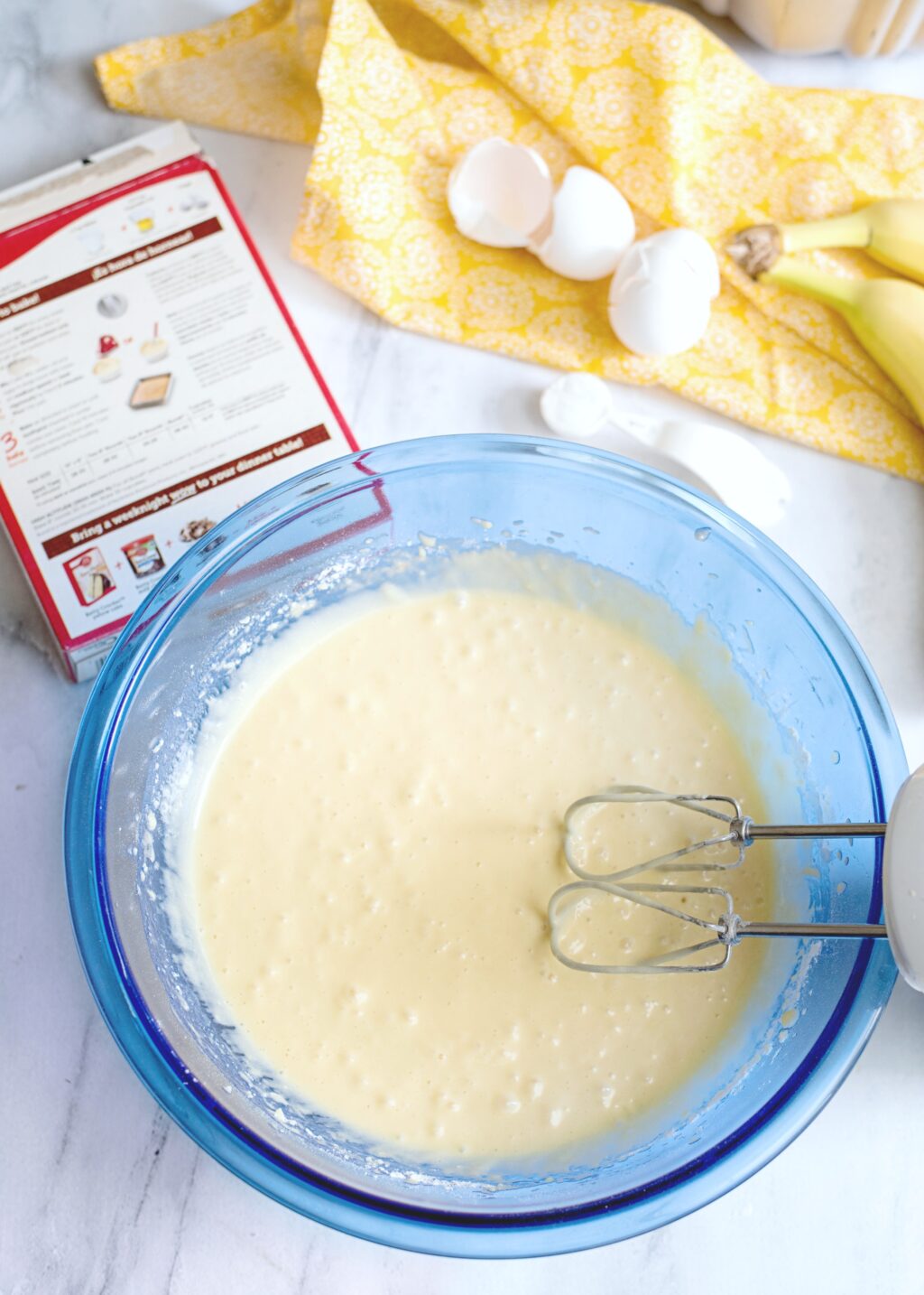Cake batter being mixed with a hand mixer. Bananas and egg shells on napkins surround it. 