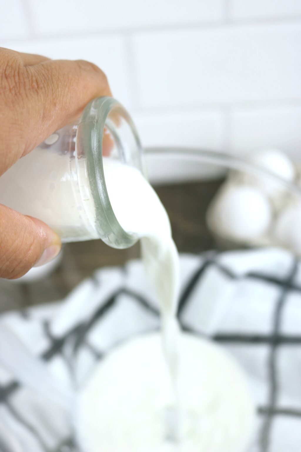 Half and half cream being poured into glass bowl. Napkin and eggs are pictured in the background. 