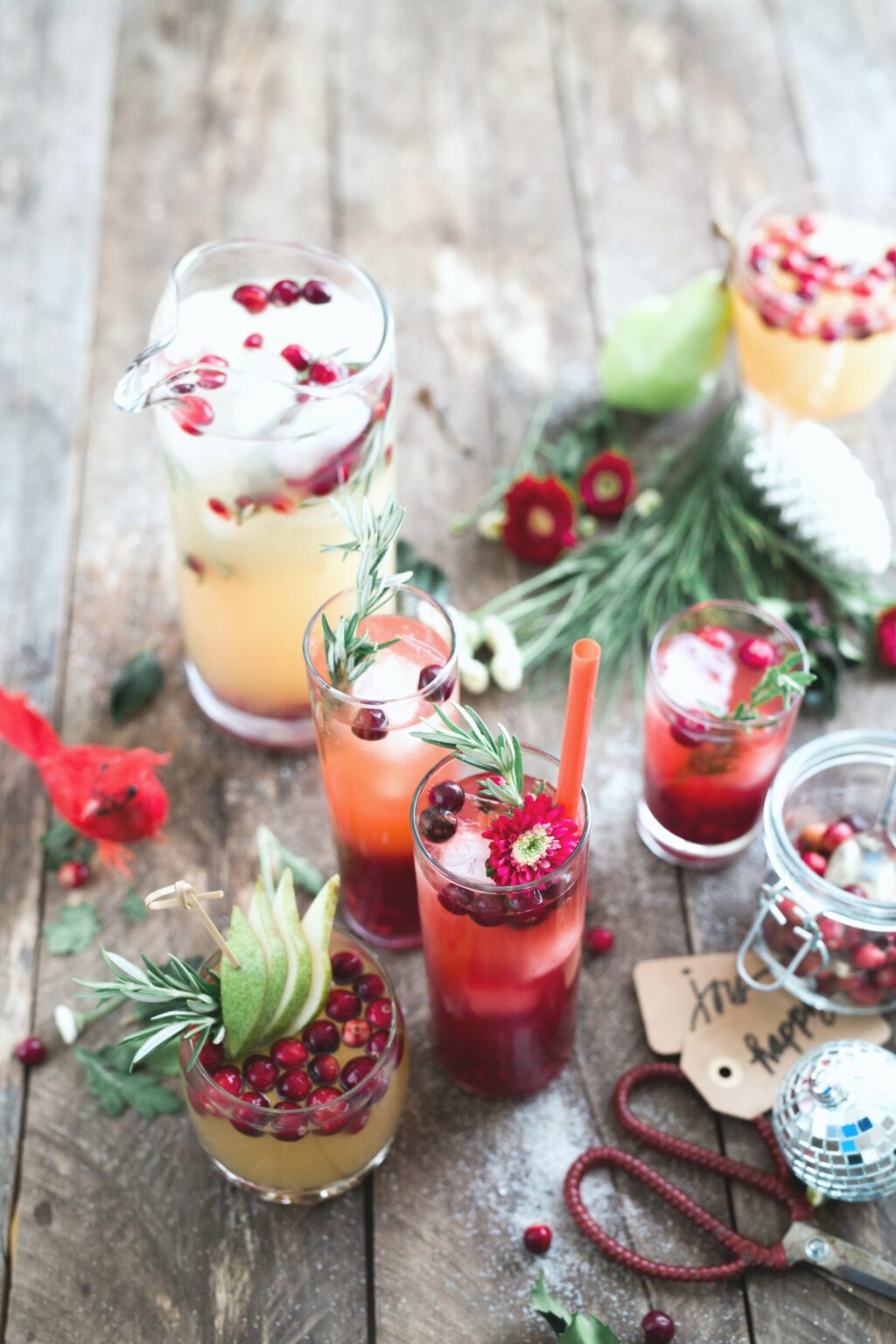 Glasses filled with beautiful cocktails and toppings that are festive for the Christmas season. Other Christmas items surround the drinks on a wooden table. 