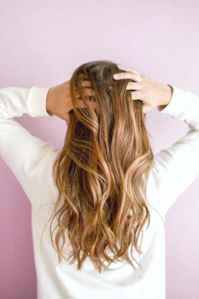 Girl stands towards a pink wall, with her hands to her head. Her hair is the main focus.