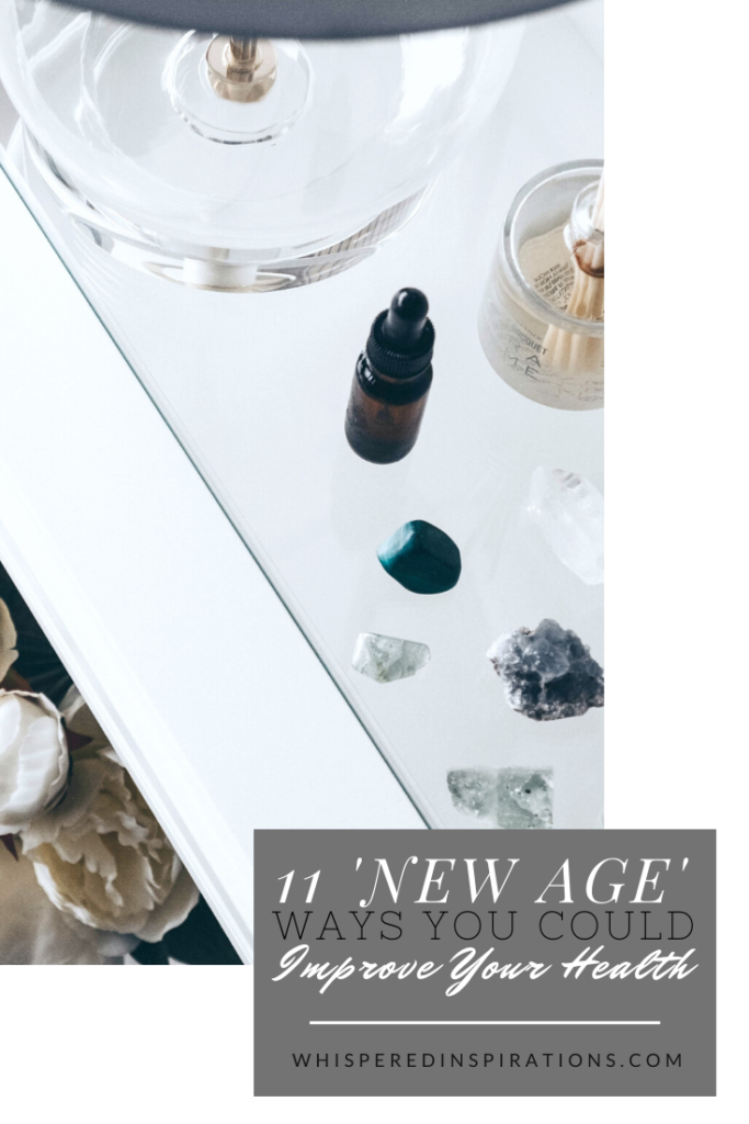 A bedroom side table is adorned with a clear lamp, white flowers, and you can see crystals, essential oils, and incense. A banner reads, "11 New Age Ways You Could Improve Your Health."