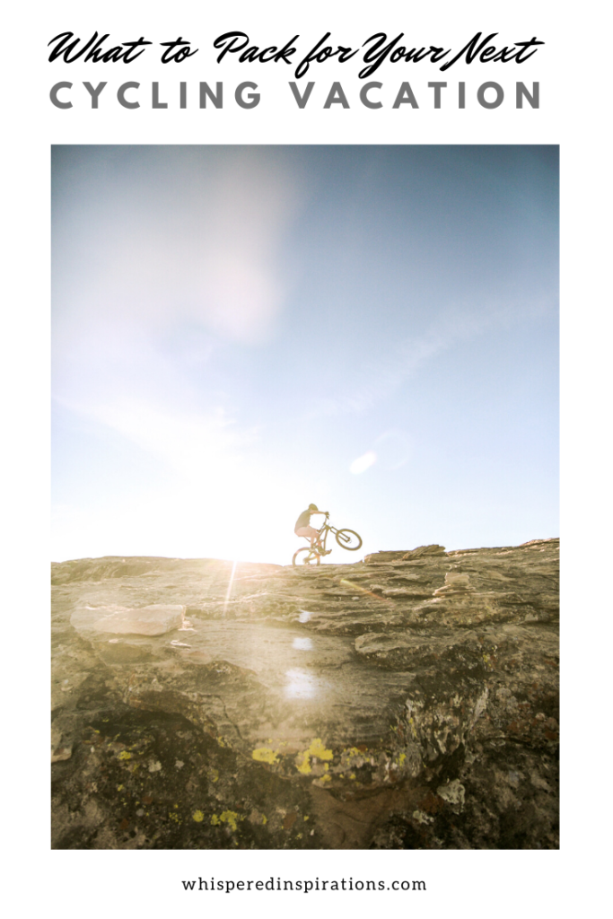 A banner reads, "What to pack for your next cycling vacation," a picture of Cyclist on top of a mountain. The sun shines brightly behind him. Have you ever thought of taking a cycling vacation? If not, why not? If you're an active and outdoorsy person, you can see your destination in a unique way. #packingtips #traveltips