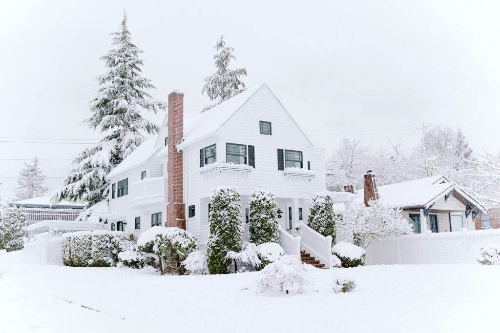A beautiful older home, covered in snow. The snow covers everything around it, including evergreen pines behind the home. A red chimney pops out in the sea of white.