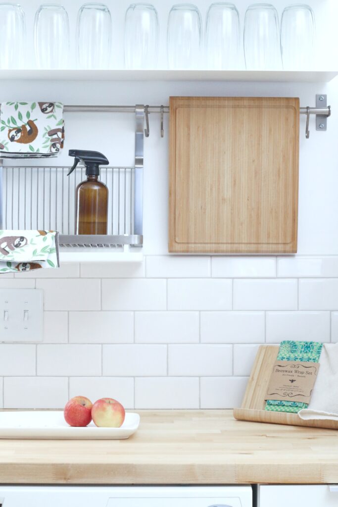 A kitchen is shown, against subway tiles, you can see a shelf with cleaning products and a cutting board. 