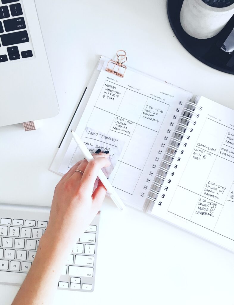 A woman uses an agenda to write down her schedule and short term goals. A computer is also seen upon the desk. 