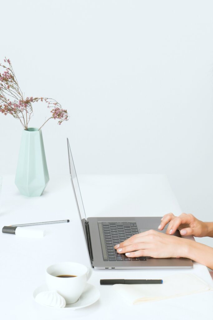 A woman sits at home with her laptop. Coffee, flowers, pens, and other office supplies are seen. A laptop is shown as an invitation to contact us.