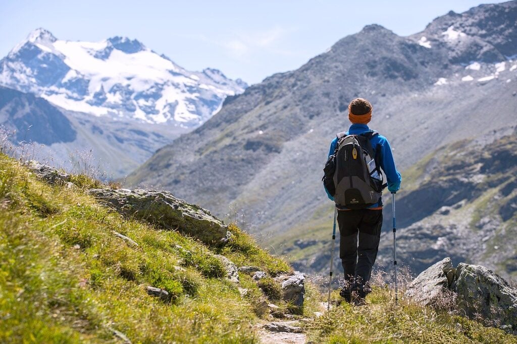 Man is geared to go hiking. The mountains are seen in front of him and he has walking sticks. 