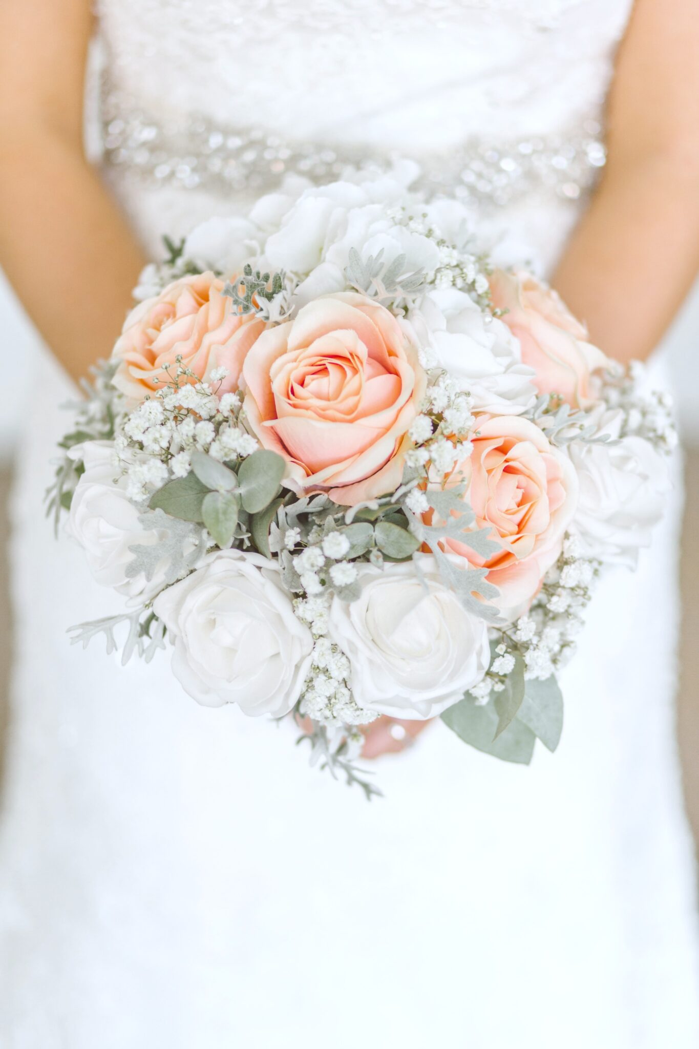 A bride holds white, coral, and beige roses in a bouquet. Her arms are only visible. 