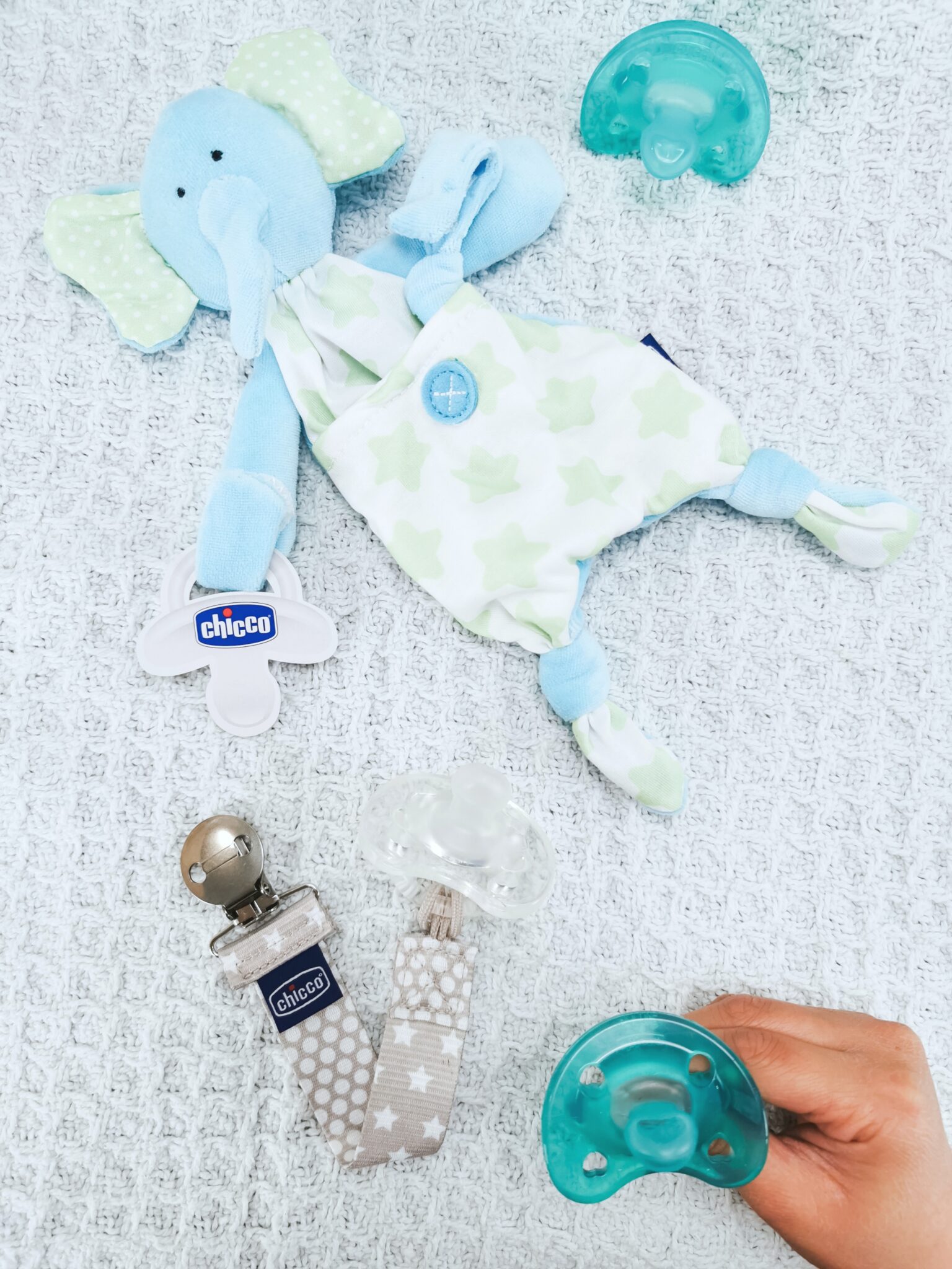 A little hand holds a Chicco pacifier on top of a blue blanket. 