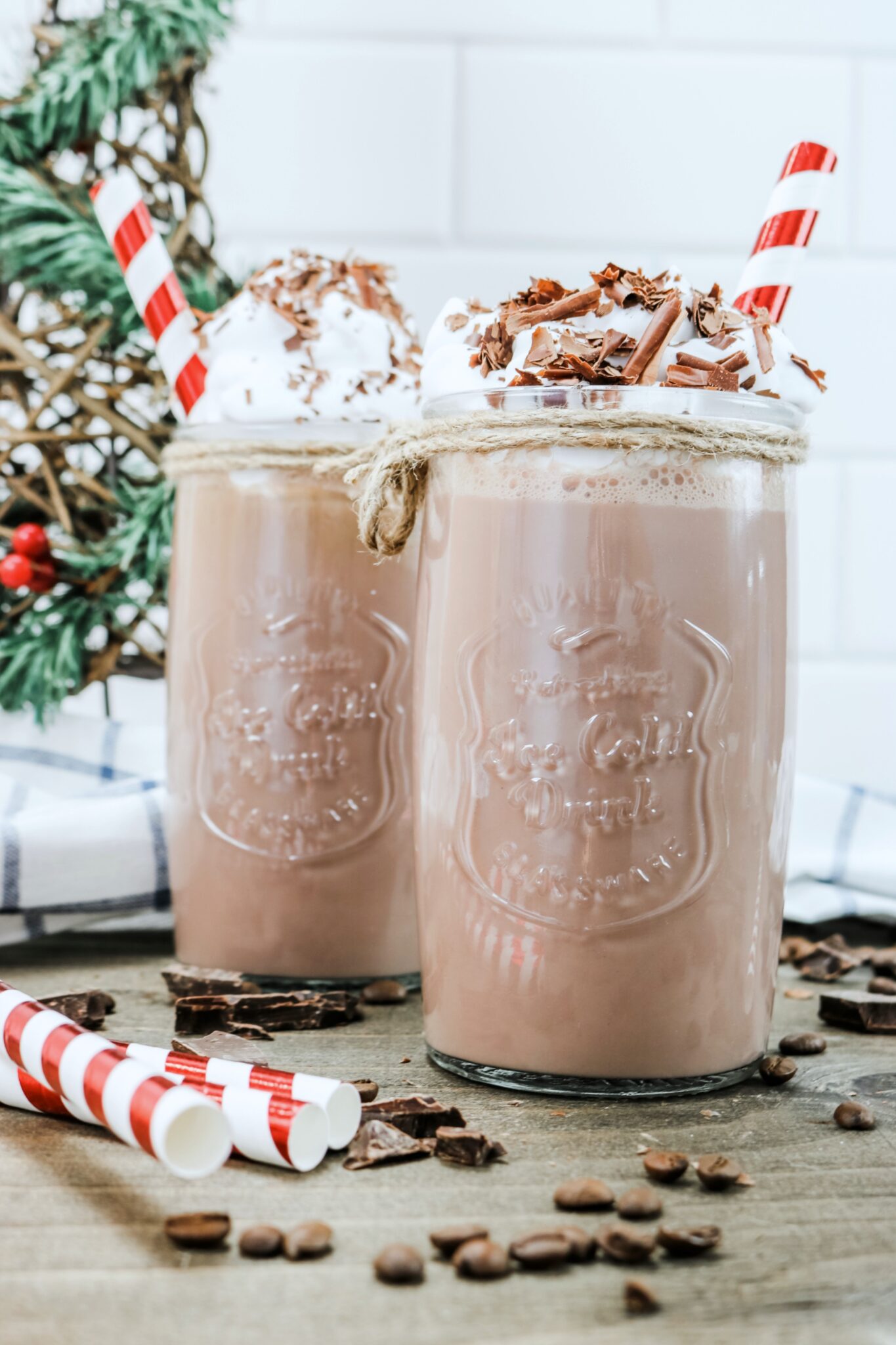 Close up of two Hazelnut Protein Iced Mocha drinks. A tree is in the background, as well as coffee beans and chocolate chunks surround them.