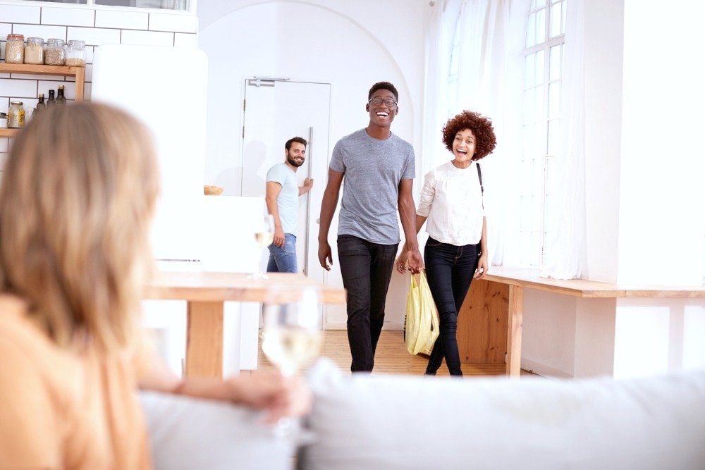 A couple walks in and happily greets their friend on the couch sipping on wine. The husband closes the door behind them.