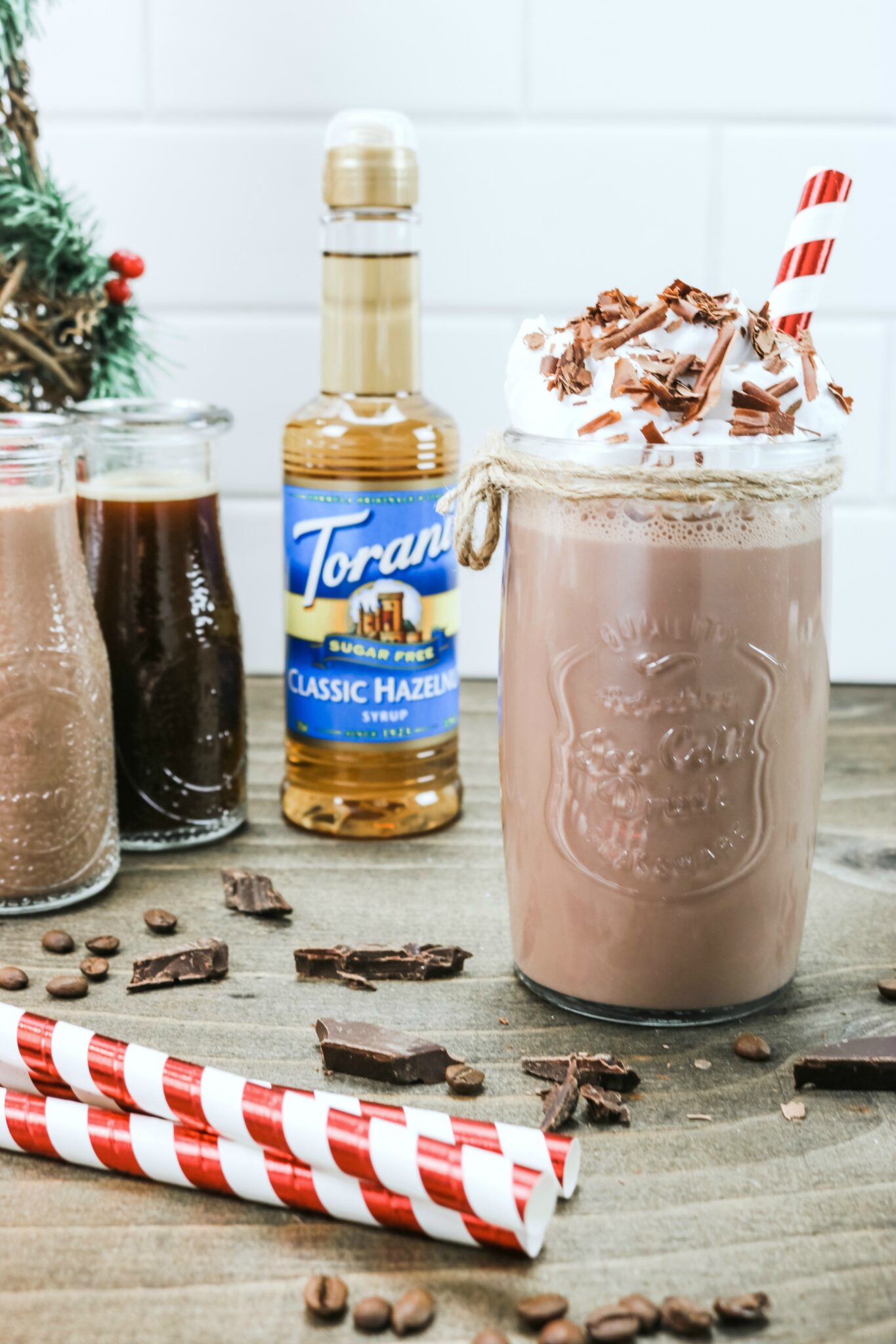 A close up of the Hazelnut Protein Iced Mocha with the Torani Classic Hazelnut Sugar Free syrup, coffe, protein shake, and chocolate are shown in the background.