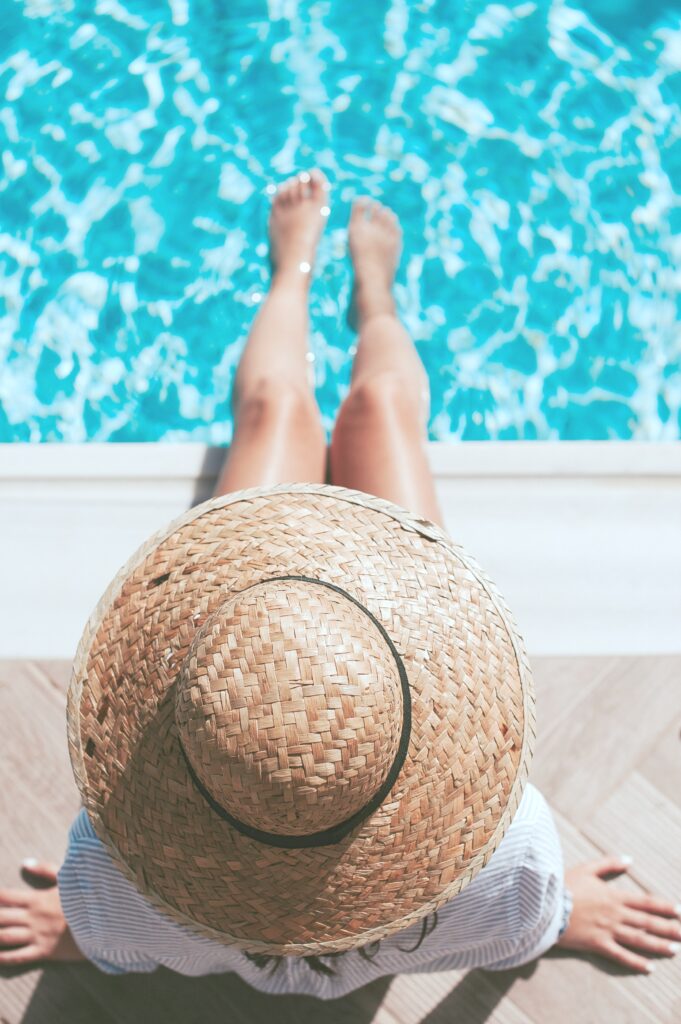 A woman sits by the edge of a pool. It's a top view of her, only her hat, and legs dipped in the pool are seen. 