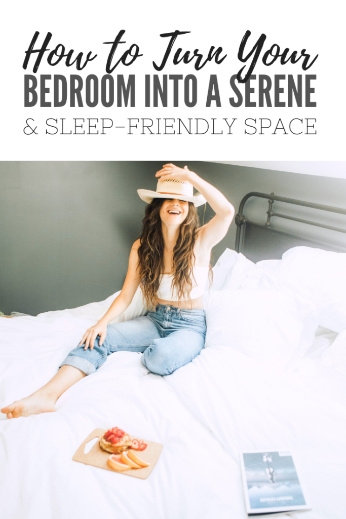 A woman smiles and holds down a hat on her head. She is in bed and has a tray of fruit and a camera on the bed. A banner reads, 'How to Turn Your Bedroom into a Serene & Sleep Friendly Space.'