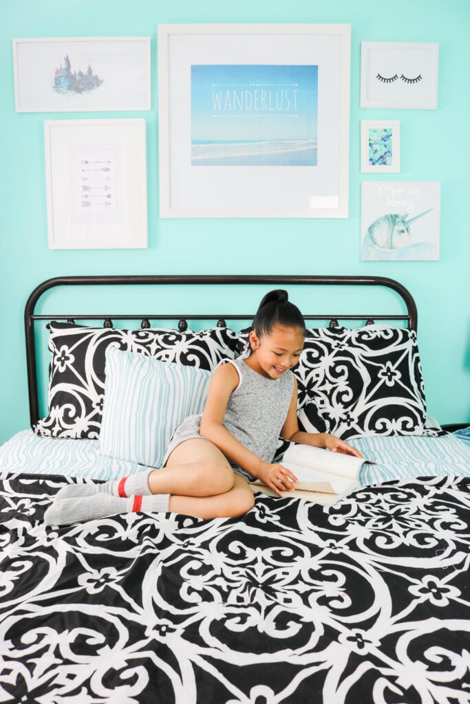 Mimi sits on the bed reading a book. Behind her is a teal wall with her travel-inspired wall art. 