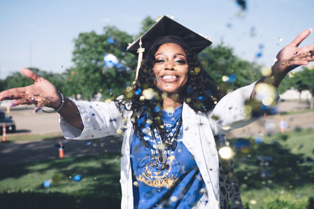 Woman is graduating after forging a career path in healthcare. She is wearing a graduation cap and a stethoscope and throwing glitter in the air.