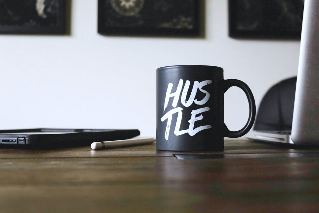 A mug with Hustle written on it sits on a wooden desk. This shows that investing can be a way to earn peace of mind. 