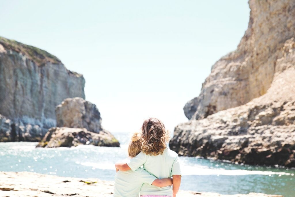 Only the backs of two young sisters are seen as they embrace. Together, they watch the waves of a beautiful seascape.