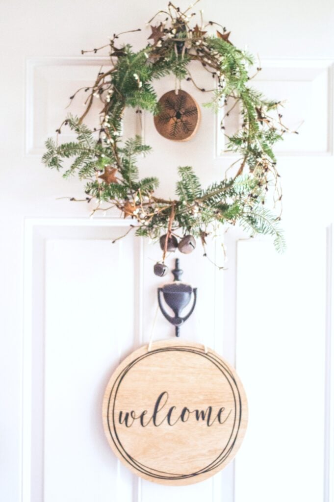 A door with a small wreath and a door knocker. Against the white door, there is a wooden welcome ornament.