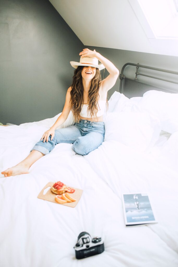 A woman smiles and holds down a hat on her head. She is in bed and has a tray of fruit and a camera on the bed. She has found a way to make her bedroom a sleep-friendly space.