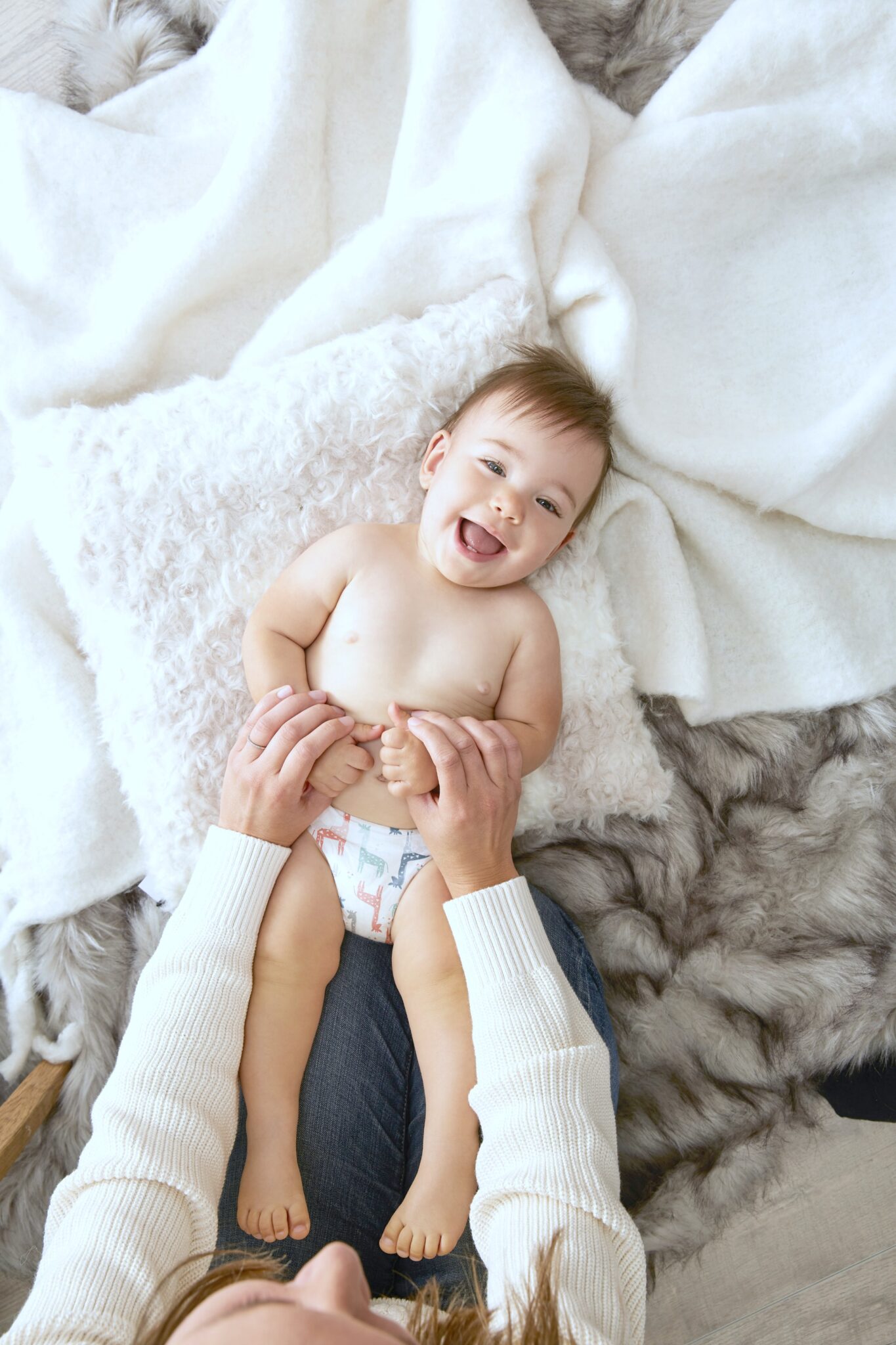A smiling baby is being tickled by his mom, it is a top view looking down at the baby. He lies on blankets.