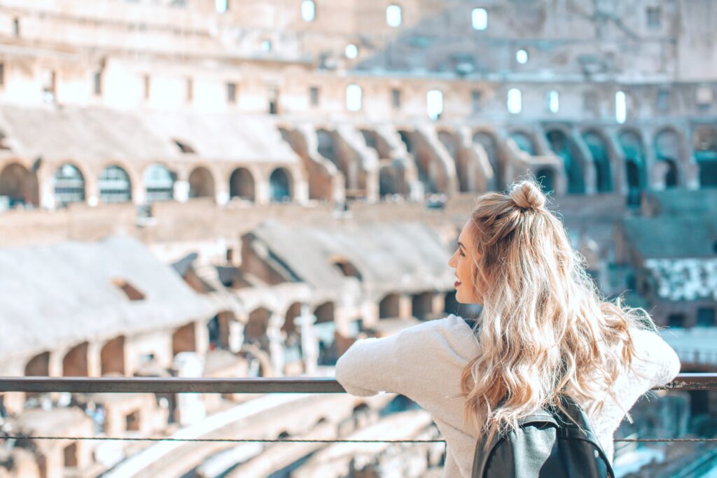 A teenager is pictured with a back back and is smiling with her head turn. In front of her, is a gorgeous stadium. She looks hopeful and happy.