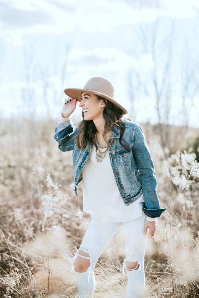 Woman stands in a field and smiles, you can see her wearing a trendy outfit and various metal accessories.