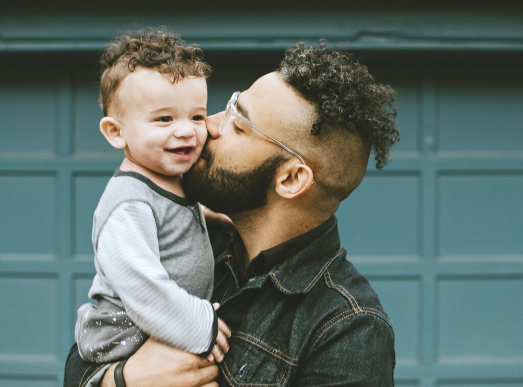 Young Dad kisses his son, against a teal garage.