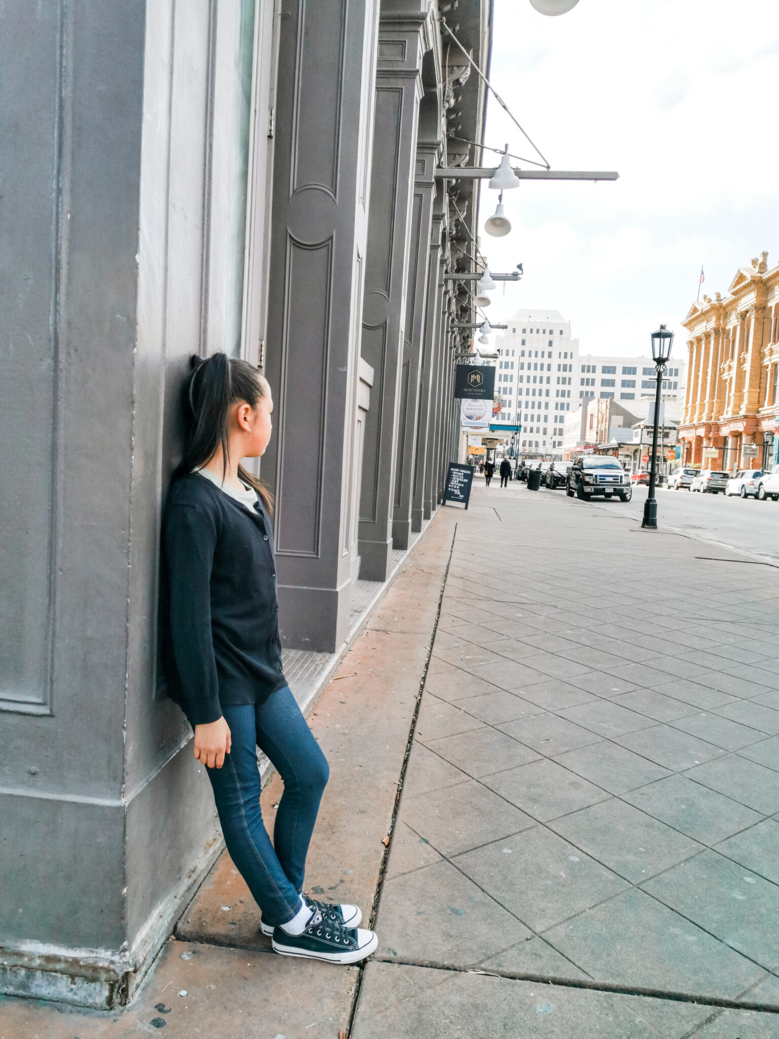 Mimi leans up against a wall by herself, the street is empty and only a few people can be seen at a distance.