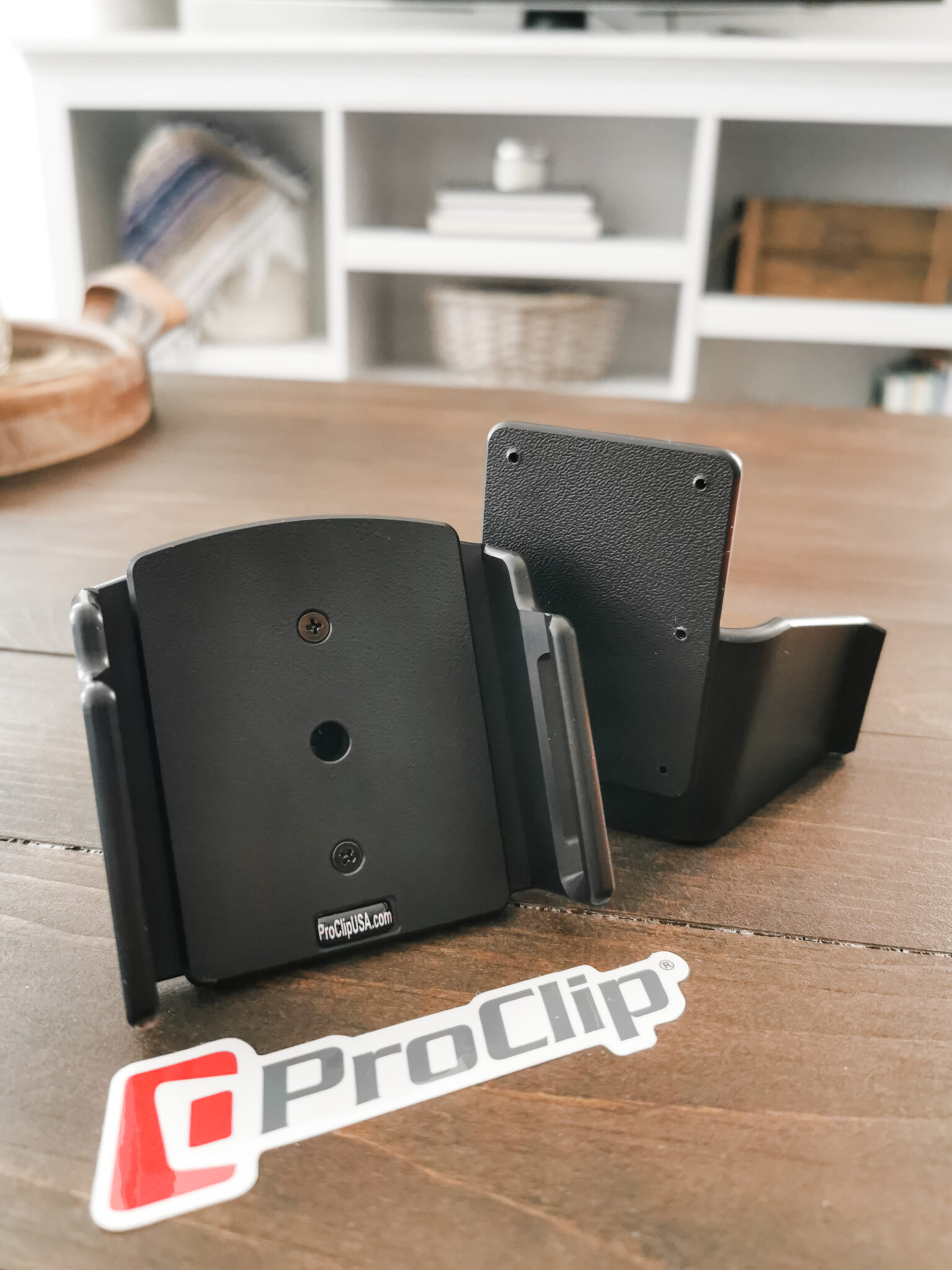 A ProClip mount and standard phone holder are shown on a wooden table. A ProClip sticker sits in front of them. A bookcase is seen in the back.